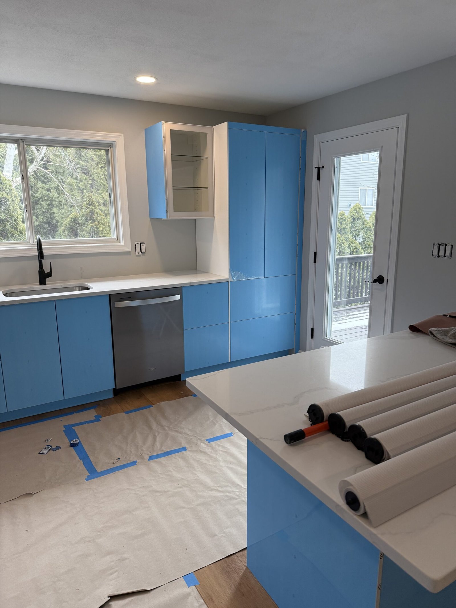 Glossy white kitchen cabinetry covered with blue protective installation film beneath a wide window in Salem Massachusetts