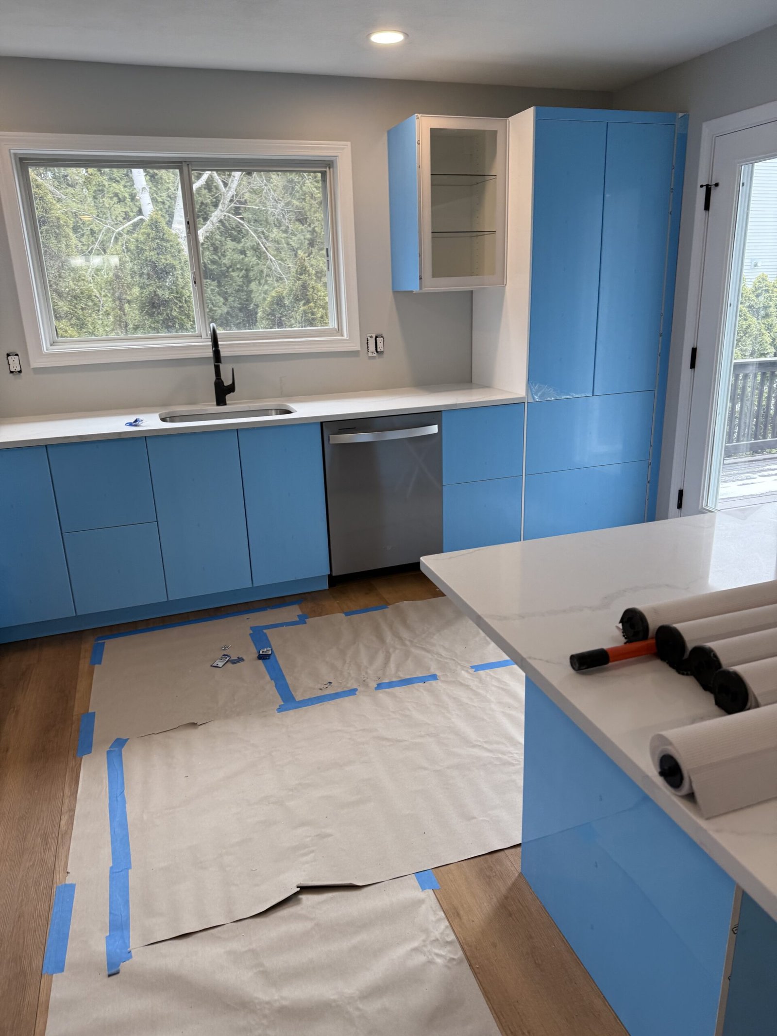 Wide view of glossy white kitchen cabinets protected by blue film, sink window, and island during a Salem kitchen remodel