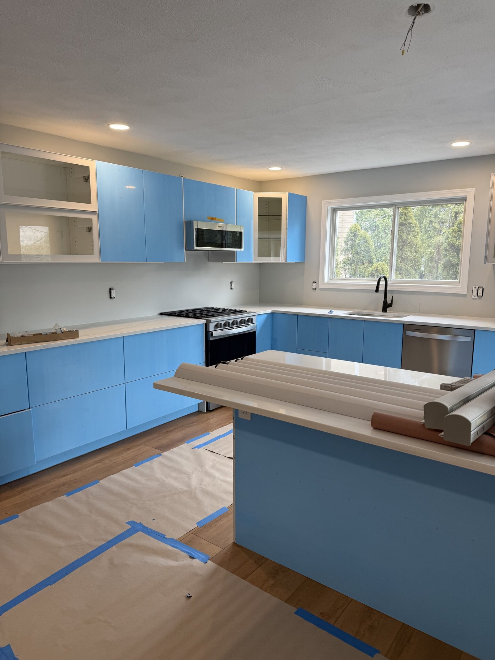 Wide near-completion view of a Salem kitchen with glossy white cabinetry under blue protective film, white counters, range wall, and island