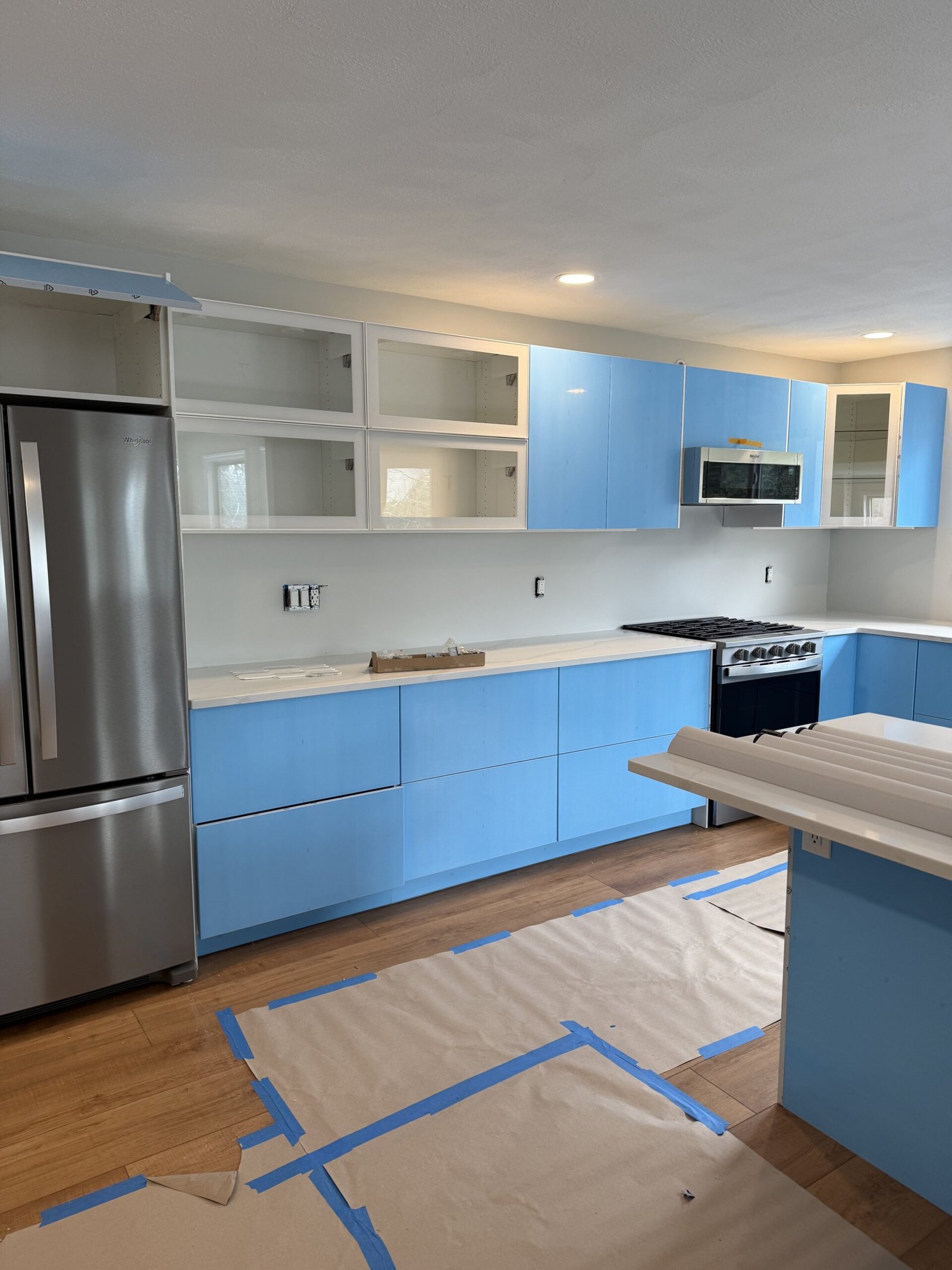 Stainless refrigerator, glossy white cabinets protected by blue installation film, range, and island during completion of a Salem kitchen renovation